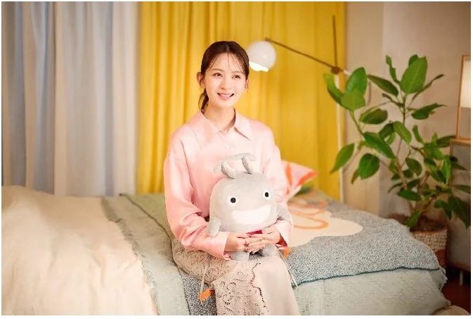 笑顔の若い女性がベッドに座り、グレーのぬいぐるみを抱えています