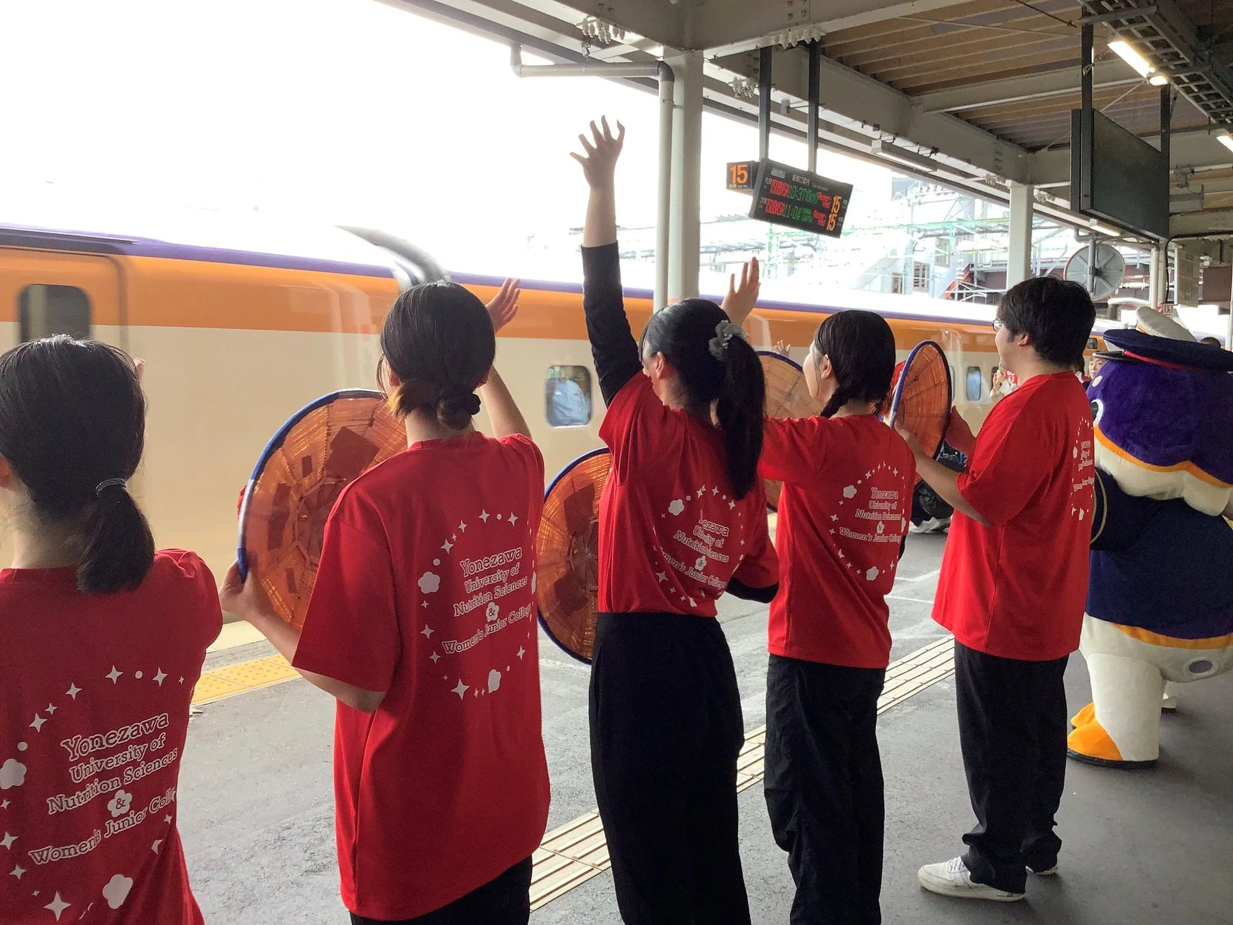 新幹線のホームで、赤いTシャツを着た学生たちが列車に向かって手を振っている様子