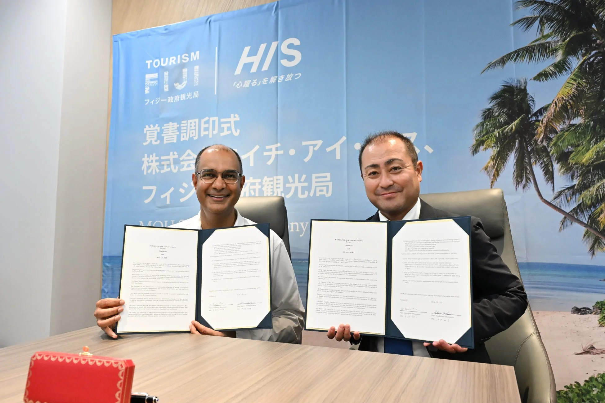 フィジー政府観光局とH.I.Sの覚書調印式で、2人の男性が笑顔で文書を掲げています。フィジー観光に関するビジネス提携を示す重要な瞬間です。