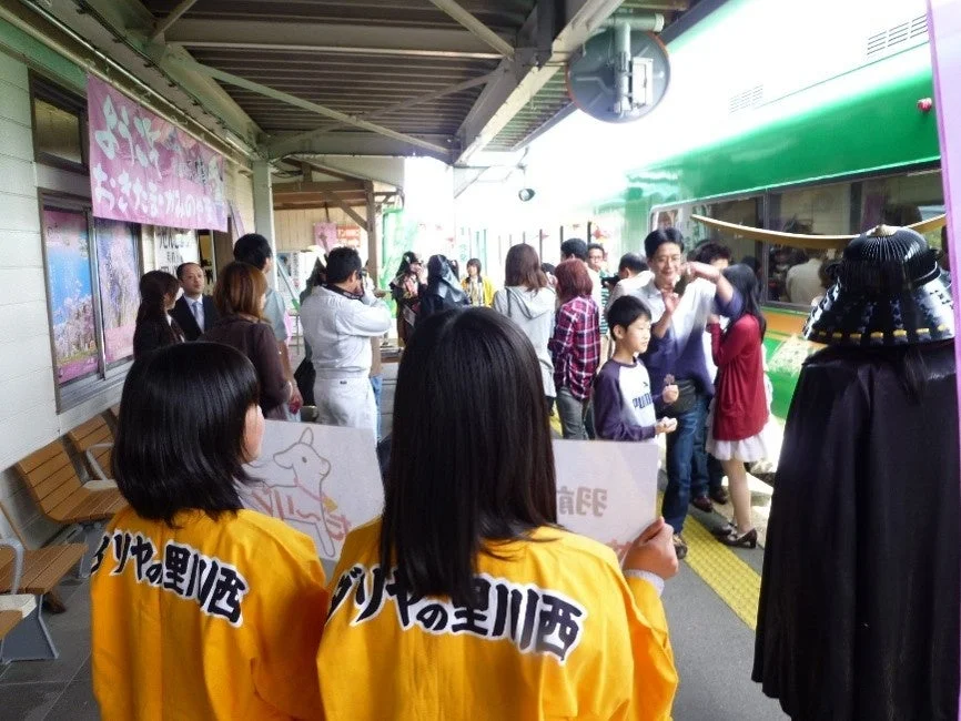 「ダリヤの里川西」と書かれた黄色いシャツの女性たちが立つ駅のホームで、緑色の列車と多くの人々が集まるイベントの様子