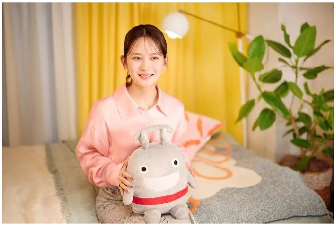 ピンクのシャツを着た若い女性が、笑顔で灰色のぬいぐるみ抱き、部屋のベッドに座っています