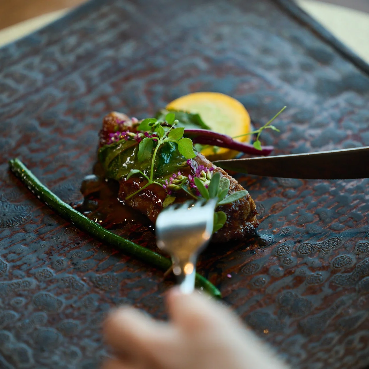 ダークな質感の皿に美しく盛り付けられた肉料理が写っています。ソースのかかった肉には新鮮なハーブと小さな花が添えられ、奥にはレモンとインゲンが彩りを添えています。食事中にフォークとナイフで切り分けられている瞬間を捉えた一枚です。