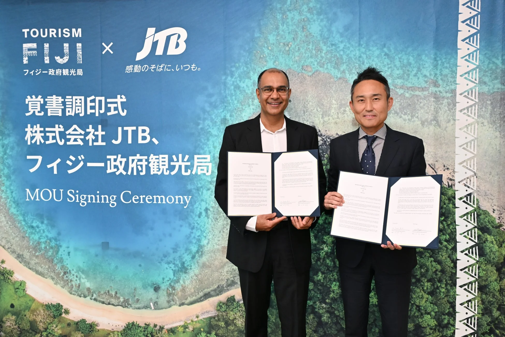 JTBとフィジー政府観光局による覚書調印式の様子です。2人の男性がそれぞれ調印された書類を持って笑顔で立っており、背景には両社のロゴと美しいビーチの風景が描かれています。