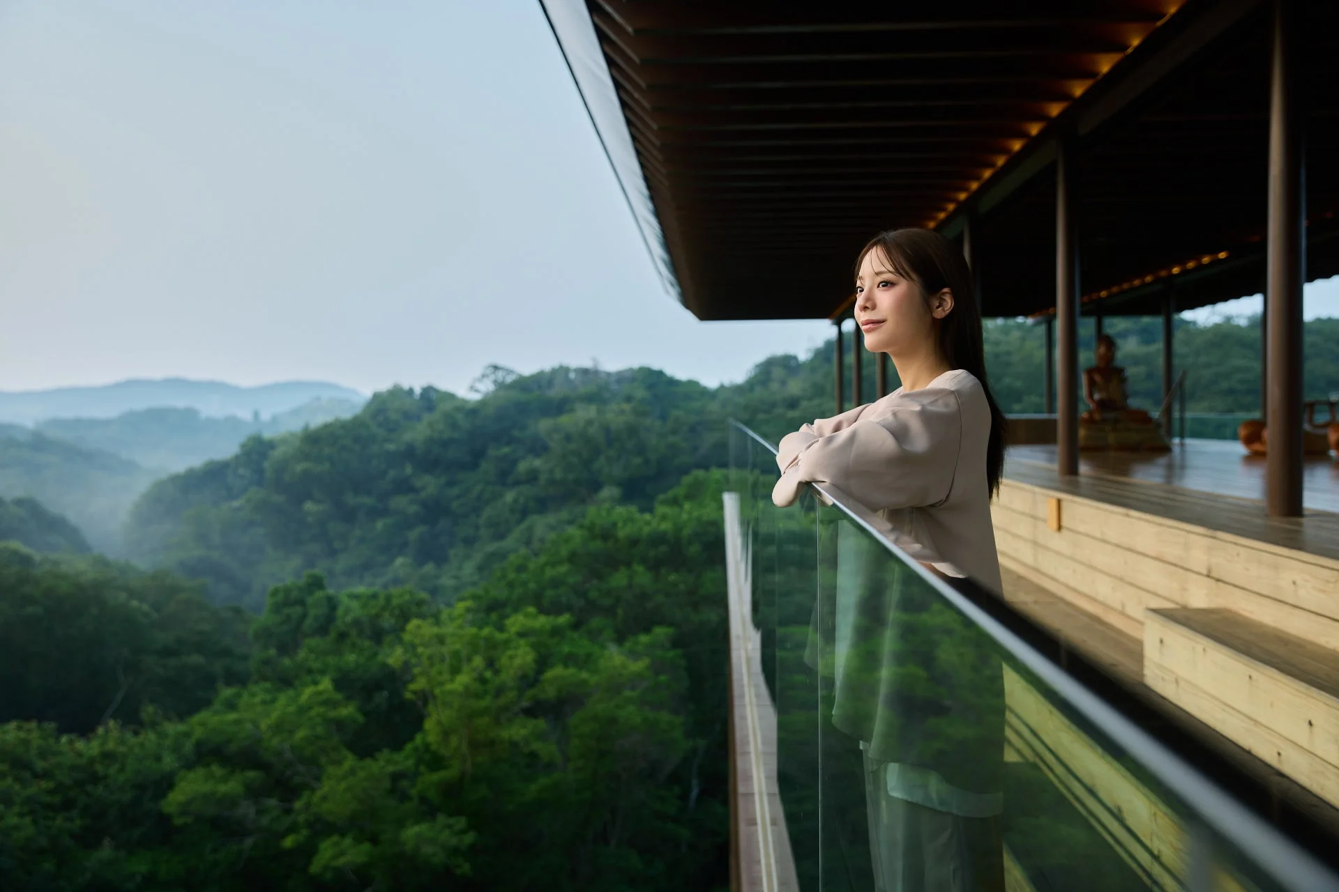 広大な緑豊かな山々を望むホテルのテラスで、若い女性が手すりに寄りかかり、穏やかな表情で景色を眺めている。背景には木造の建物と仏像が見え、リラックスしたひとときを過ごしている様子がうかがえる。