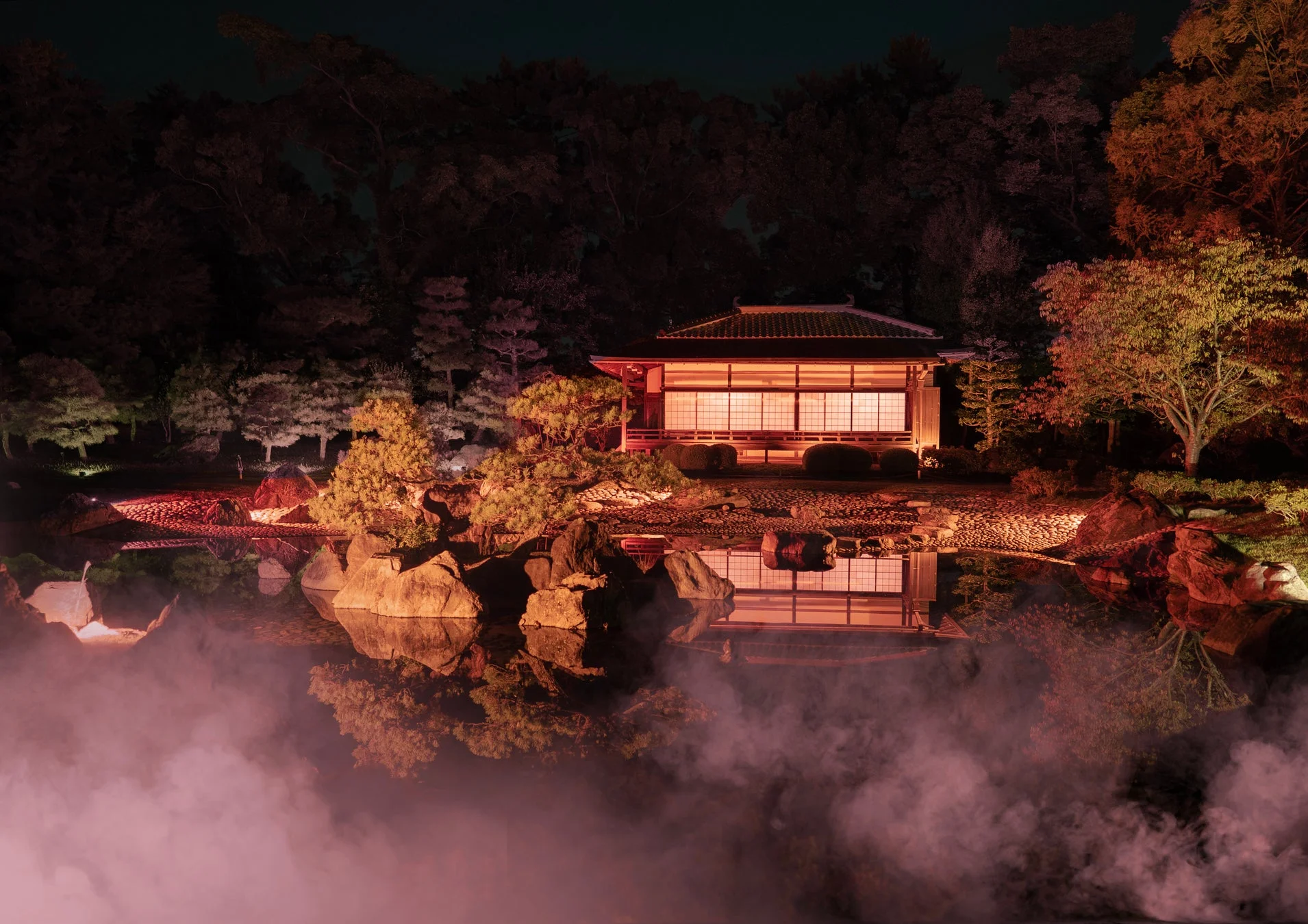 夜の日本庭園に佇む伝統的な家屋が美しくライトアップされ、池の水面に幻想的に映り込んでいます。
