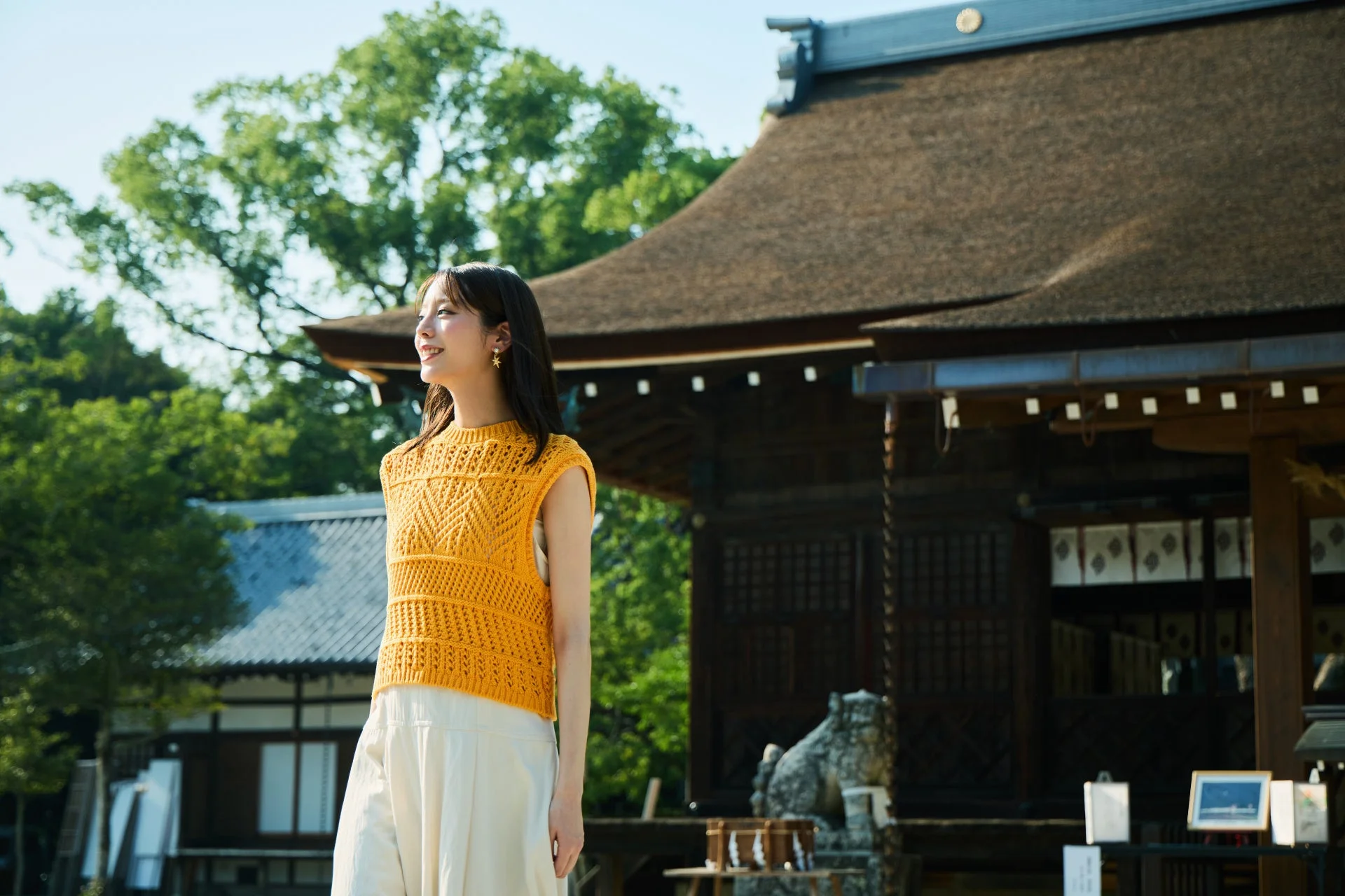伝統的な日本家屋を背景に、黄色のサマーニットを着た若い女性が笑顔で遠くを見つめる写真。晴れた日の屋外で、和風の趣が感じられる。