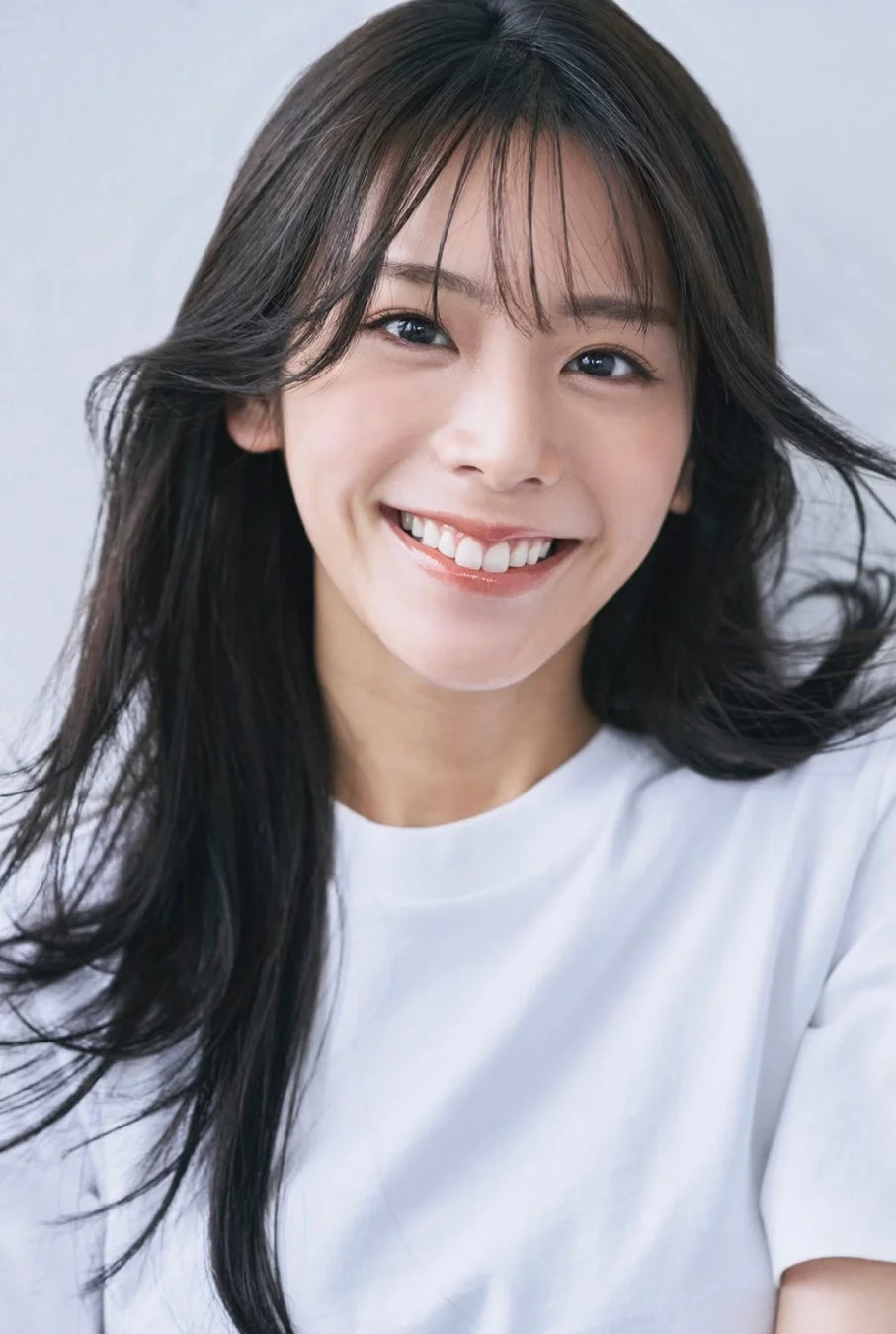 白いTシャツを着た若いアジア人女性が、カメラに向かって満面の笑顔を見せています。長く流れるような黒髪が特徴的で、明るく親しみやすい雰囲気を醸し出しています。背景はシンプルで、彼女の魅力が際立つポートレートです。