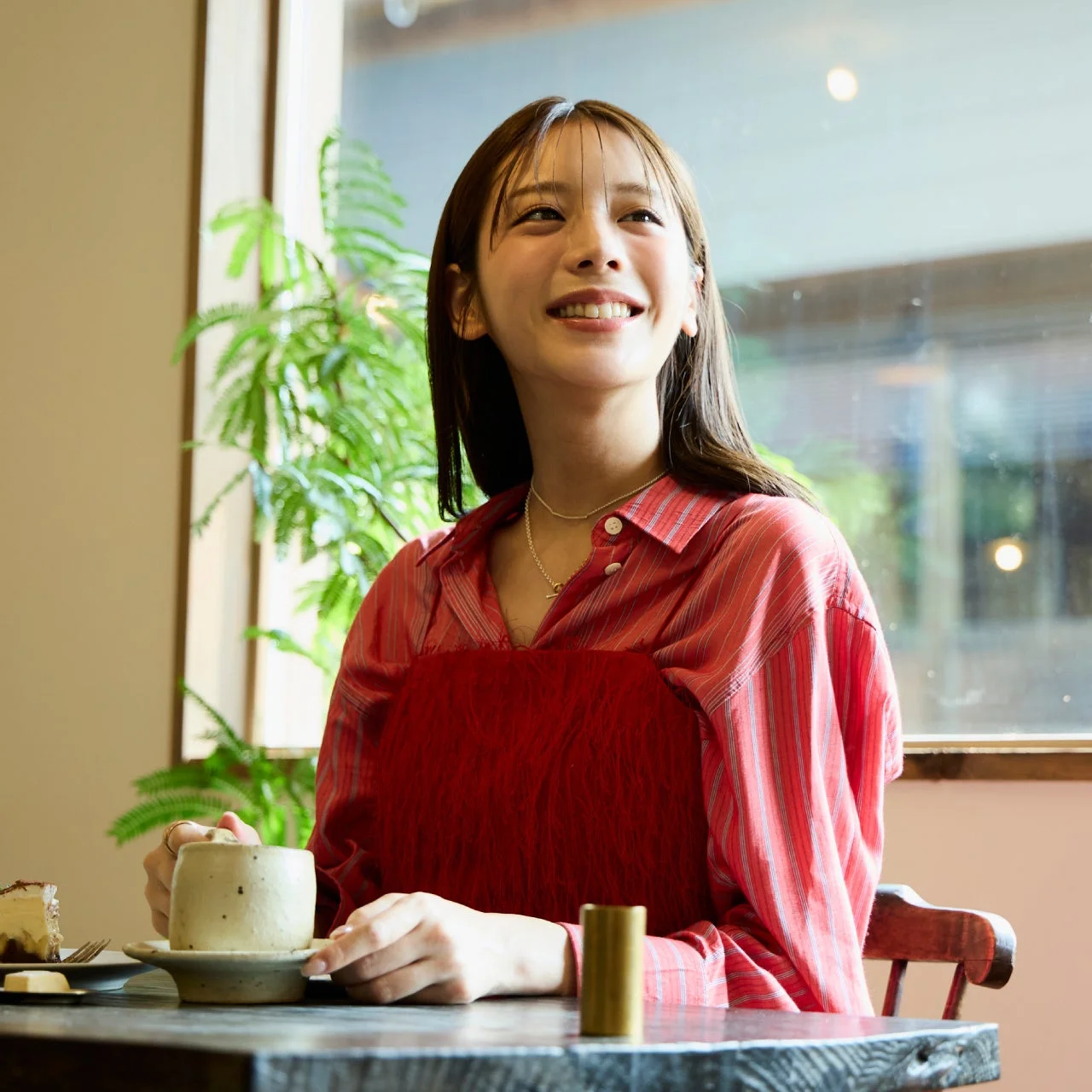 カフェでくつろぐ笑顔の女性が、カップを手に窓の外を見つめている。テーブルにはケーキもあり、穏やかな時間が流れている様子が伺える。