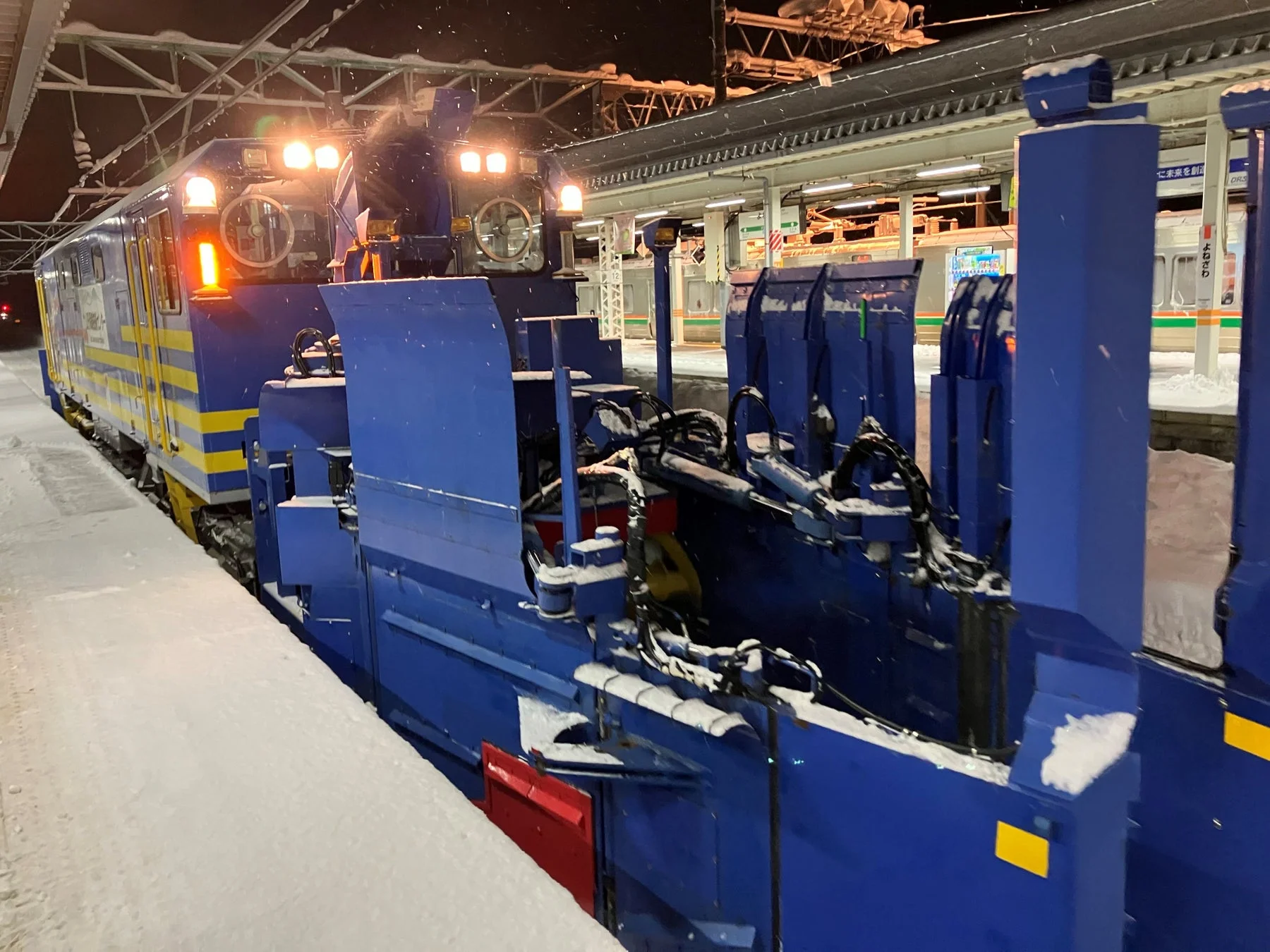 夜の雪降る駅のホームに、力強いヘッドライトを灯した青と黄色の除雪列車